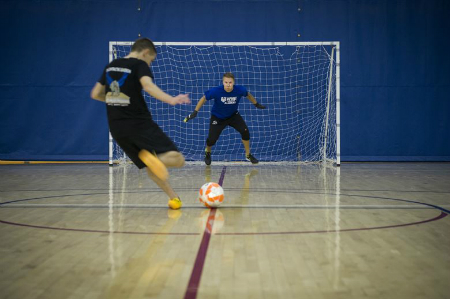 A futsal player shoots the ball at the goal guarded by a goalie