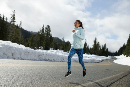 A woman running on the side of the road while the area around her is surrounded by trees and snow